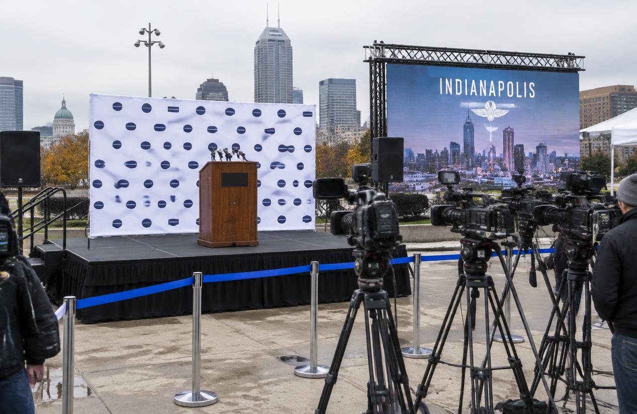 Indianapolis press conference setup with podium and microphones on a black stage, branded backdrop, LED video wall, camera row and silver stanchions with blue tape